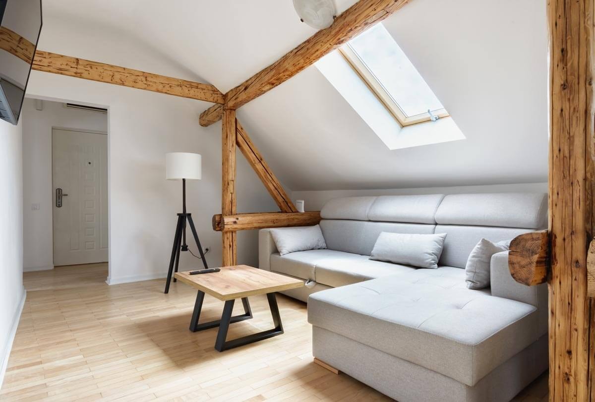 Light upholstered seating group between load-bearing wooden roof beams beneath a sloping ceiling with a skylight, opposite a wall-mounted TV.