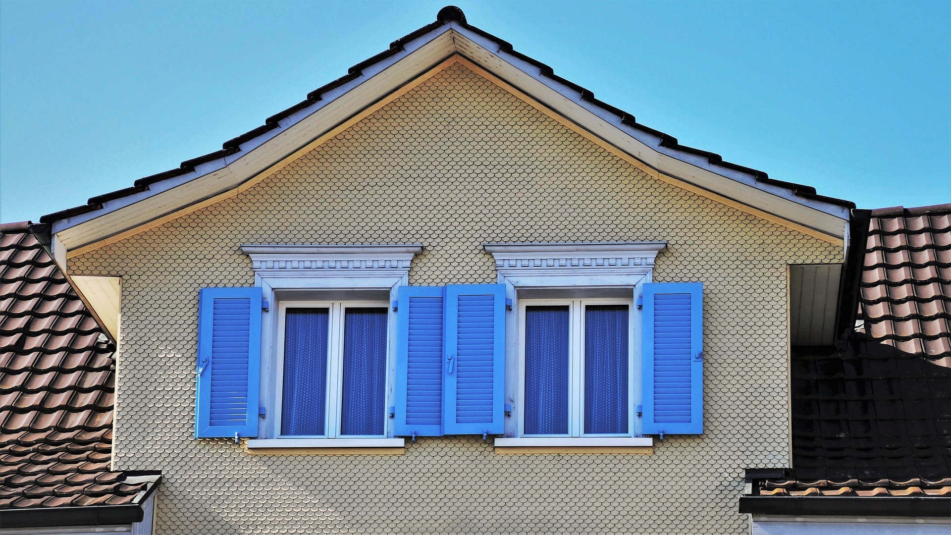 Close-up of a dormer window with two windows and blue shutters, beige wall render and reddish-brown roof shingles against a blue sky.