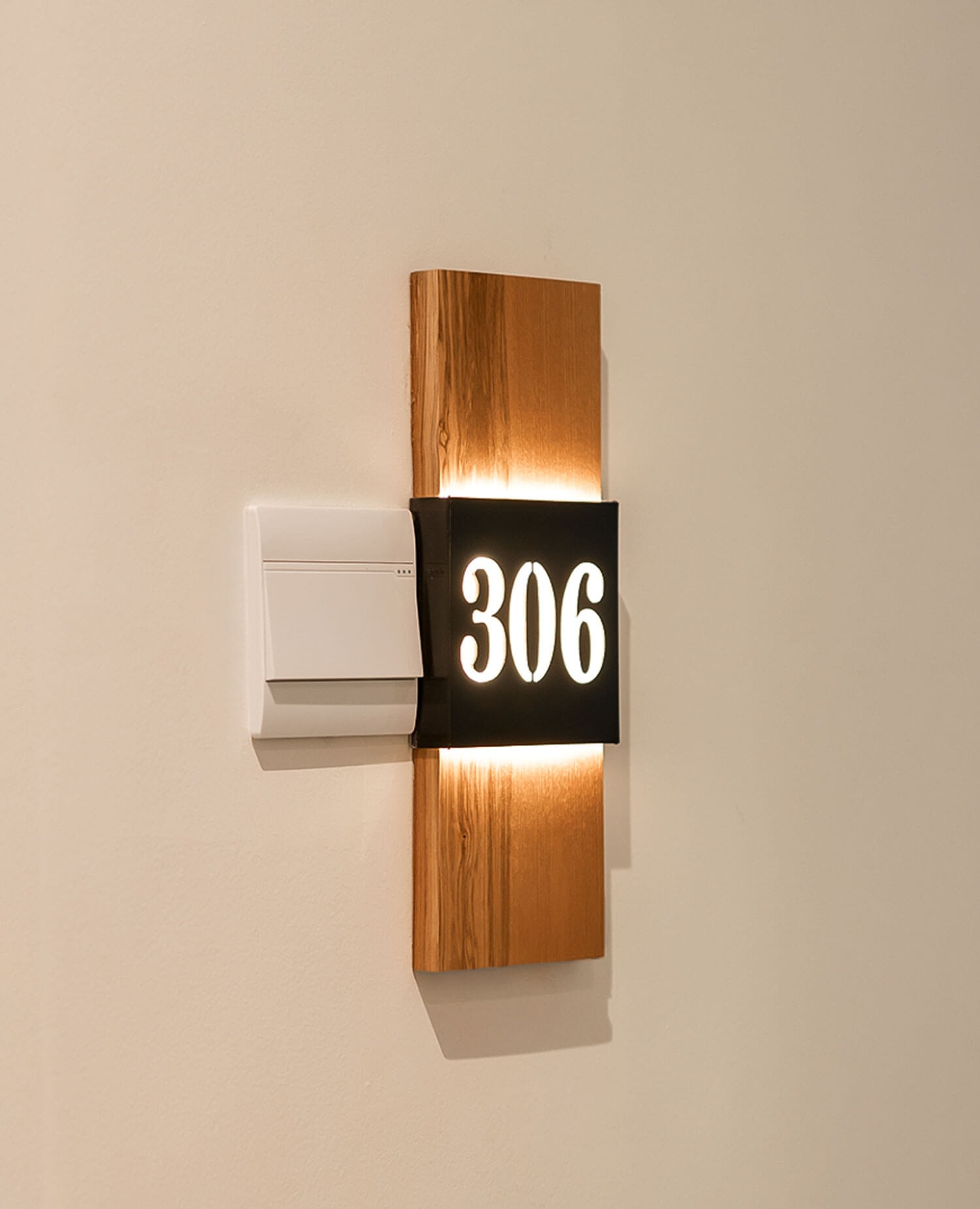 Close-up of an illuminated room number '306' mounted on a wooden backing on the wall, with a light switch next to it