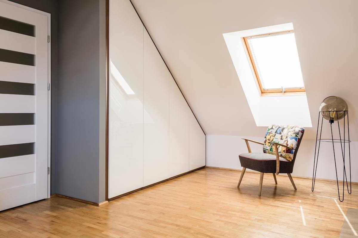 View of the corner of a room with a sloping ceiling, with an armchair and a lamp beneath a skylight and a bedroom door next to a built-in wardrobe on the left.