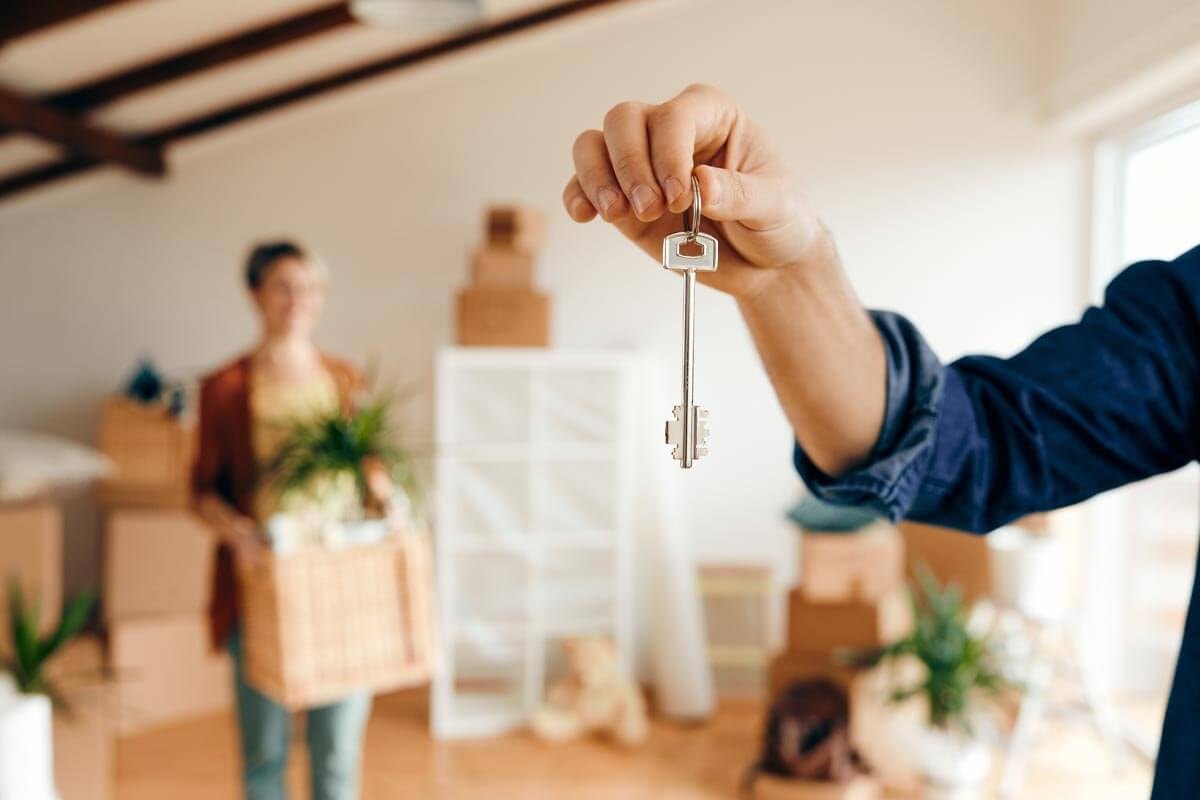 Close-up of an arm holding a house key towards the camera; in the background a woman with moving boxes in an empty apartment is vaguely visible.